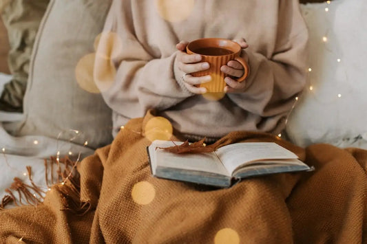 Kobieta siedzi pod kocem i trzyma ciepły kubek w szaro-beżowym swetrze w jesiennym klimacie